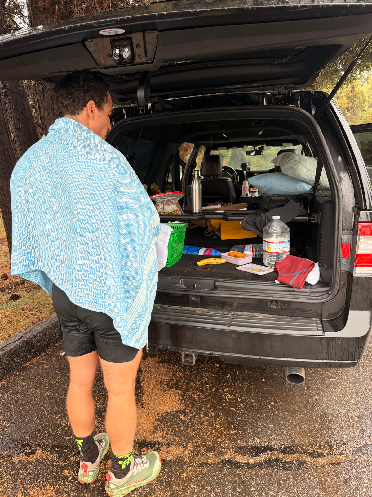A man wrapped in a light blue towel stands with his back to the open trunk of a black SUV, which is loaded with camping gear and supplies.