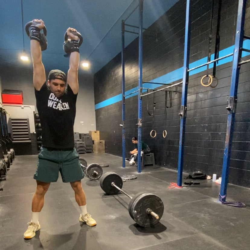 A man in a gym wearing a black graphic t-shirt, green shorts, and white sneakers performs a weightlifting exercise with a barbell on the floor. The gym has dark walls with blue lighting and various fitness equipment.