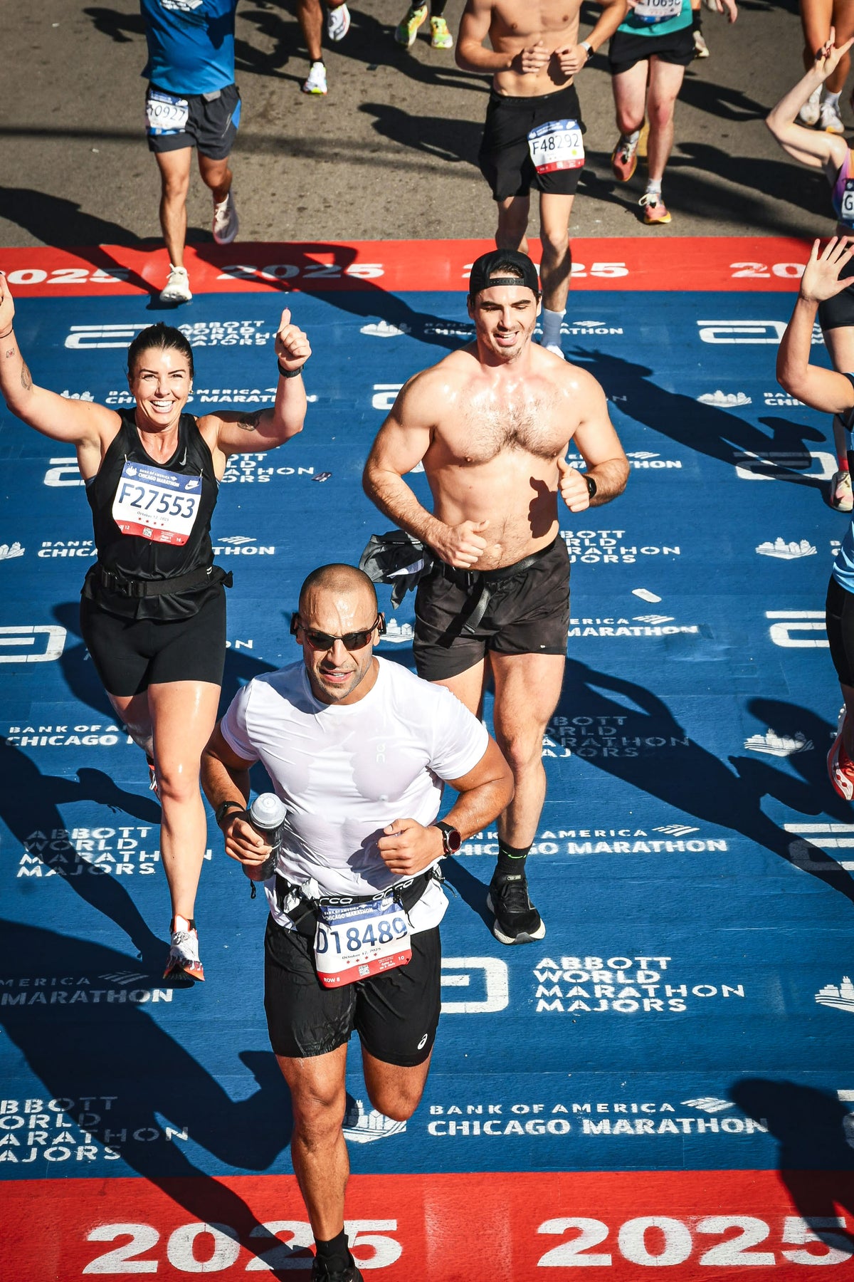 A group of marathon runners crossing the finish line on a blue branded mat with red borders. The central runner in the foreground wears a white t-shirt, black shorts, and sunglasses, with a race bib attached.